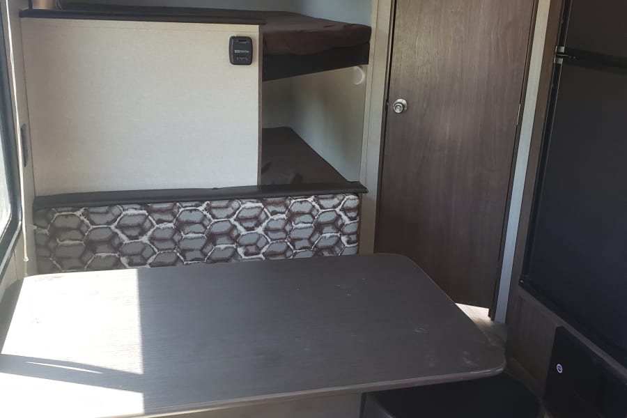 RV rental interior showing a dinette with a dark table, geometric patterned bench, bunk beds, and a brown wooden door.