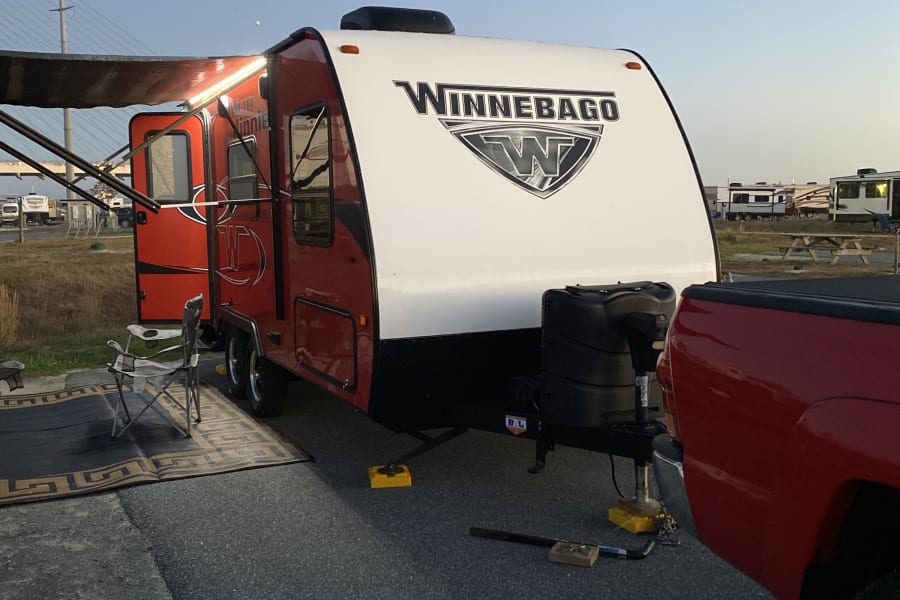 Red and white Winnebago RV rental, hitched to a truck and set up at a campsite with an extended lighted awning.