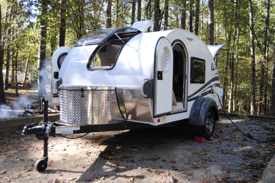 This modern teardrop RV, perfect for an **RV rental**, is set up at a wooded campsite with smoke from a fire and another camper.