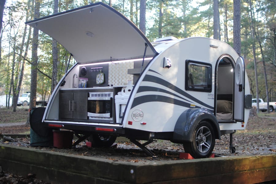 This compact teardrop camper, ideal for an RV rental, displays its open rear kitchen and open side door in a wooded campground.