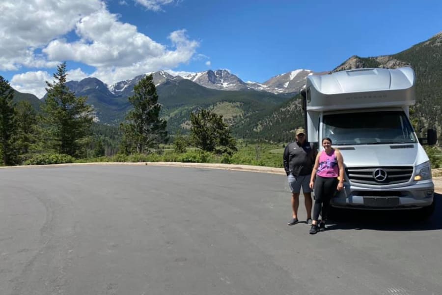 Scenic RV rental journey! A couple and their Winnebago camper van in a mountain overlook with snow-capped peaks.