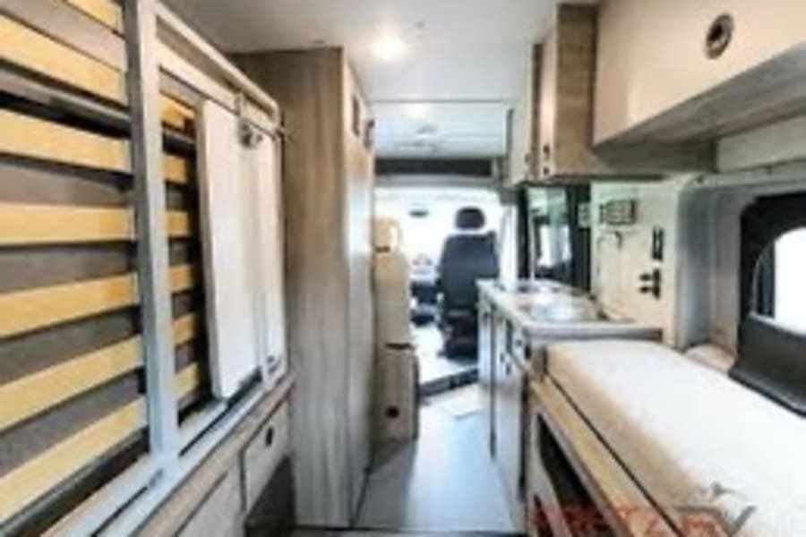 View inside an RV rental: a narrow aisle, compact kitchen, bed, and driver's area beyond. Modern camper van interior for travel.