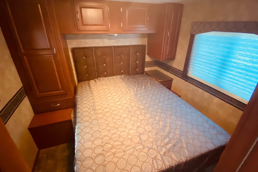 Inside an RV rental, a compact bedroom features a comfortable bed, wooden storage cabinets, and a window with blue blinds.