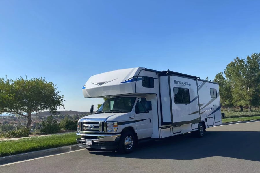 White and blue Sunseeker motorhome RV rental, parked on a sunlit road with a clear blue sky, green trees, and distant hills.