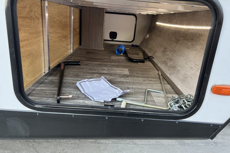 Inside an RV rental's spacious storage compartment with tools, chains, and a manual on the floor. LED-lit interior.