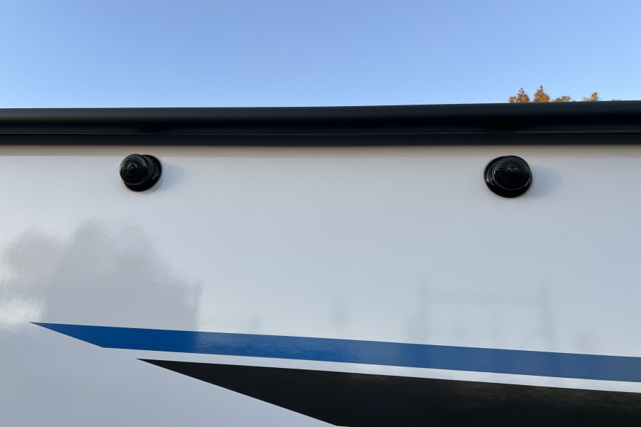 White RV rental side with black awning, outdoor speakers, and blue/black graphics. Clear sky above.