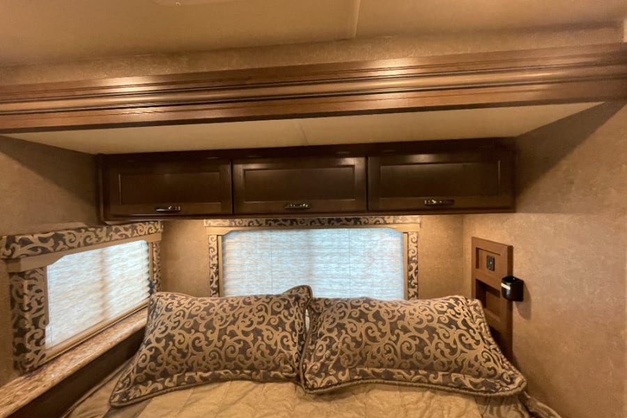 Well-appointed RV rental bedroom with dark wood overhead storage, patterned pillows on the bed, and dual windows with blinds.
