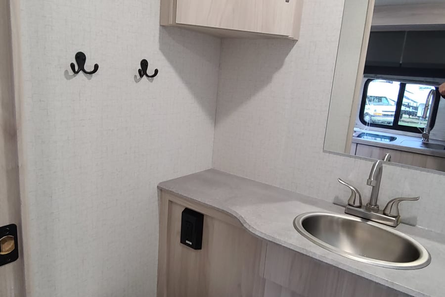 This compact RV rental bathroom features a toilet, sink vanity, overhead cabinet, and hooks for convenience.