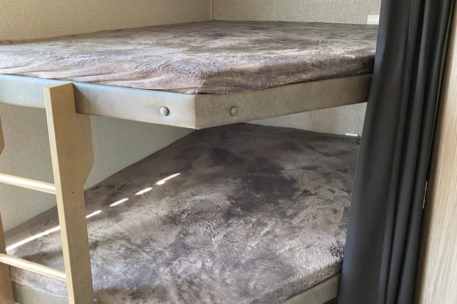 Inside an RV rental, two bunk beds with soft grey bedding, a wood ladder, and black privacy curtains create a cozy sleeping area.