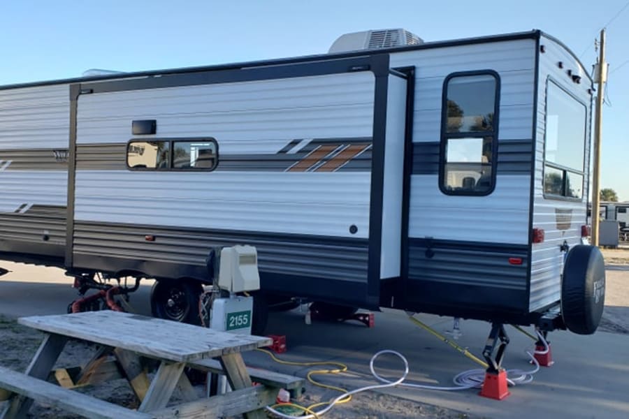 A large RV rental with a slide-out, stabilized at a sunny campground site, with a picnic table in the foreground.