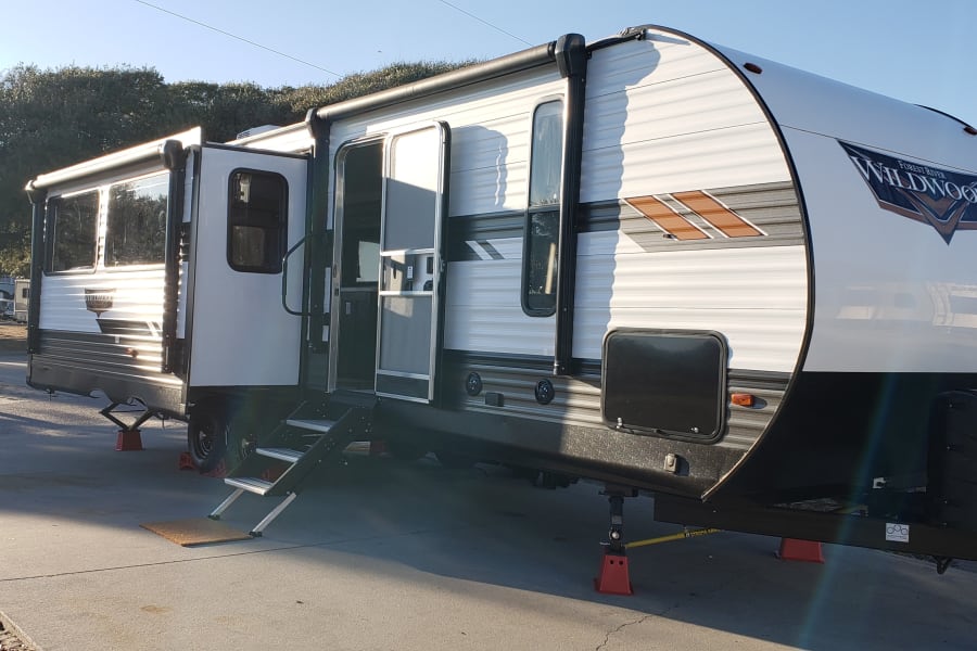 A white & black Forest River Wildwood travel trailer, ideal for an RV rental, is parked on a concrete pad at a sunny campsite.