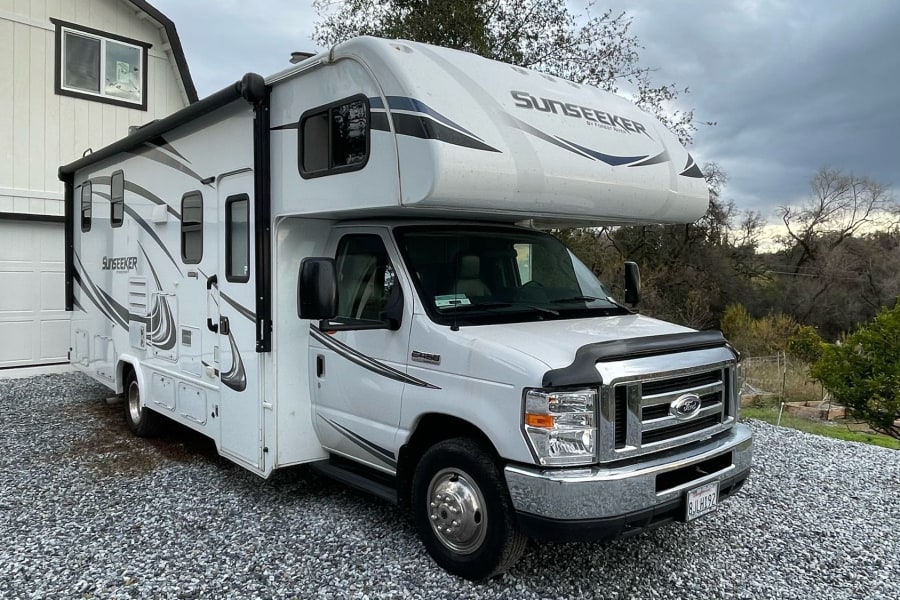 White Class C RV rental parked on gravel near a building and trees under a cloudy sky, ideal for travel.
