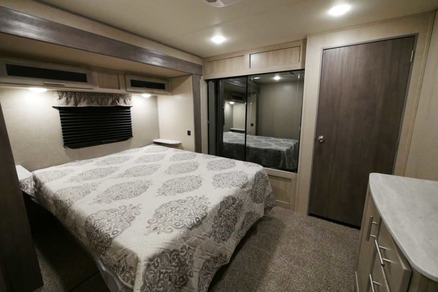Modern RV rental bedroom featuring a bed with patterned quilt, mirrored wardrobe, dresser, and window.