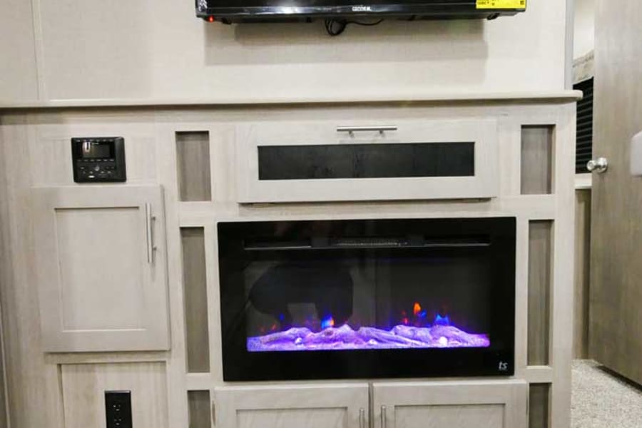 Modern RV rental living area featuring a wall-mounted TV, electric fireplace with colorful flames, and built-in storage cabinets.