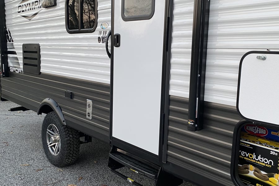 Exterior view of an Olympia Sport travel trailer, ideal for rv rental. Shows the side door, wheel, and open storage.