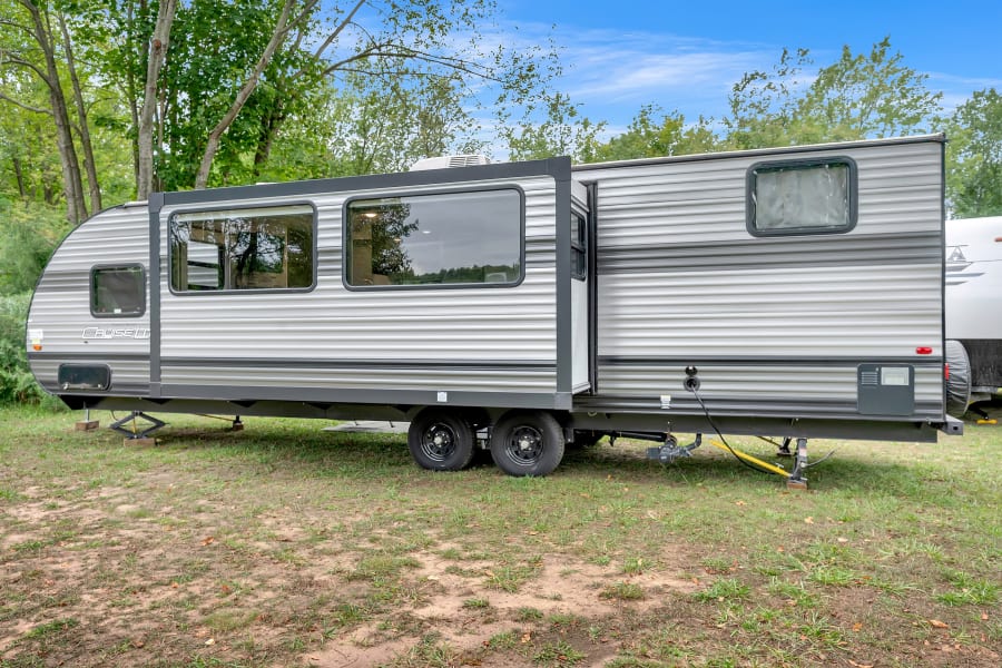 This gray travel trailer RV rental, with its slide-out extended, is set up at a leafy campsite with utility connections.