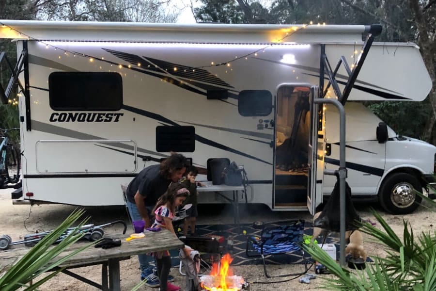 Family enjoys a warm campfire outside their spacious RV rental motorhome at a peaceful campsite.