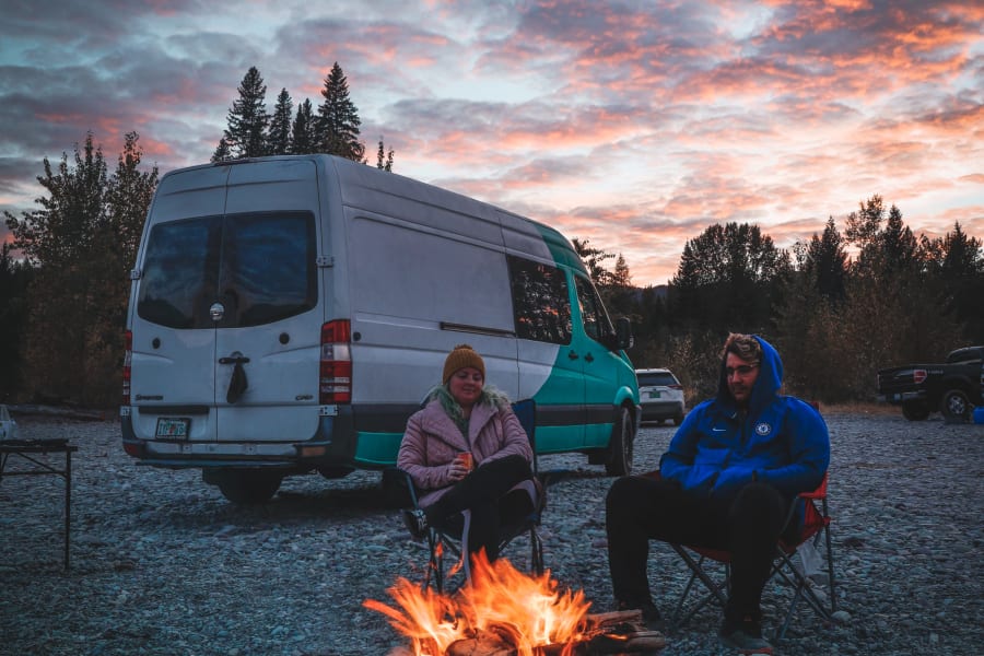 Two people relaxing by a warm campfire at sunset, beside their **RV rental** van on a gravel riverbed. Ideal for nature trips.