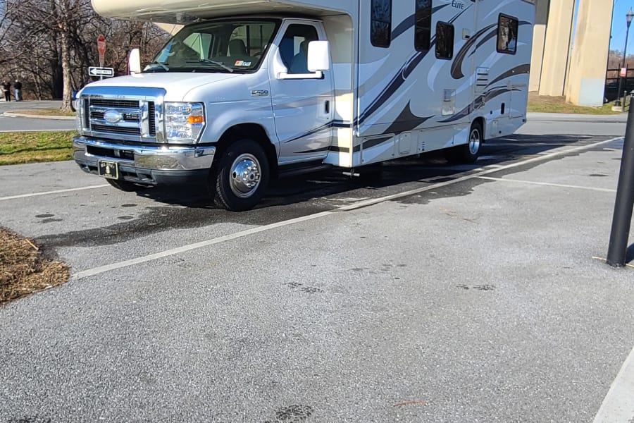 A gleaming white Ford Freedom Elite Class C RV rental is parked on asphalt with a vast, arched concrete bridge behind it.
