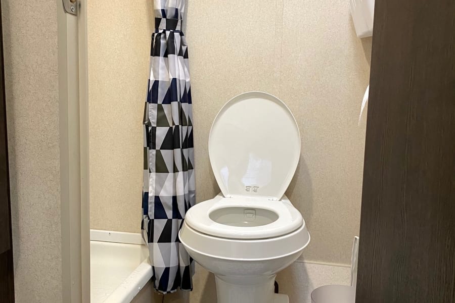 RV rental bathroom with white toilet, modern geometric shower curtain, and a blue patterned rug by the shower stall.