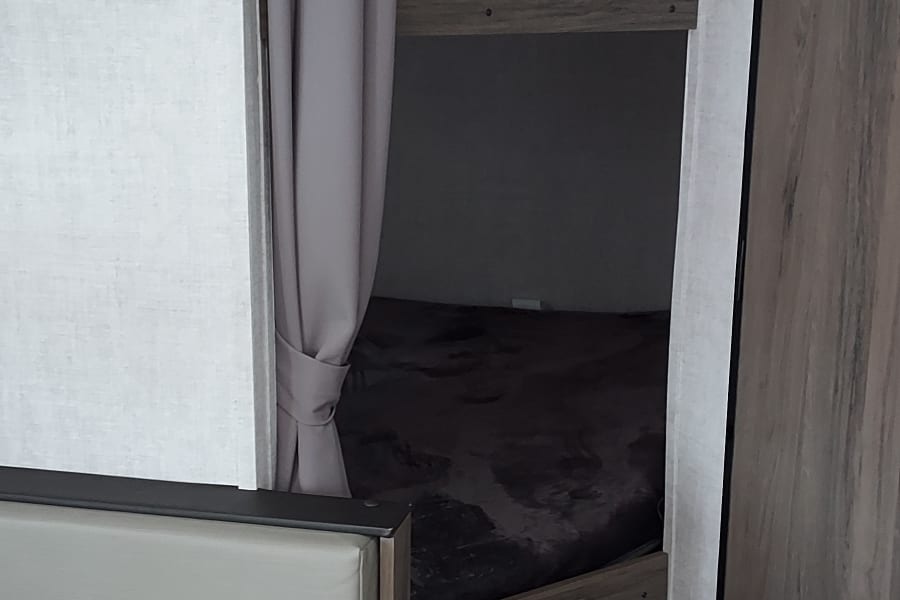 Inside an RV rental: bunk bed with gray curtain, dinette, and wood-look finishes, offering a cozy space for travel.