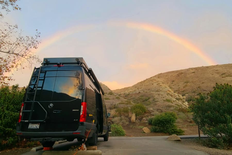 A black camper van sits under a vibrant rainbow in scenic mountains, an ideal RV rental experience.
