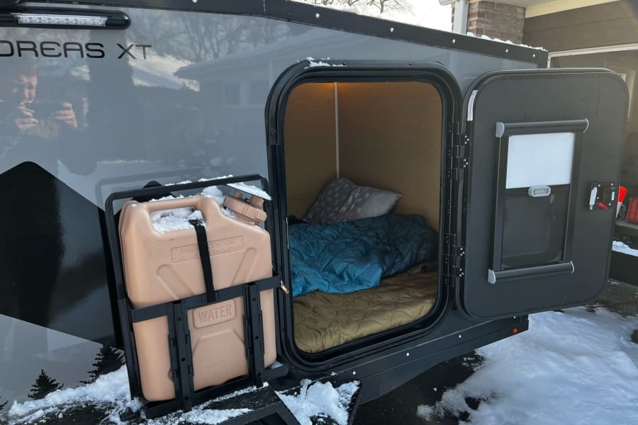 Compact rv rental with open side showing cozy bed and water storage, snow-covered for winter adventures.