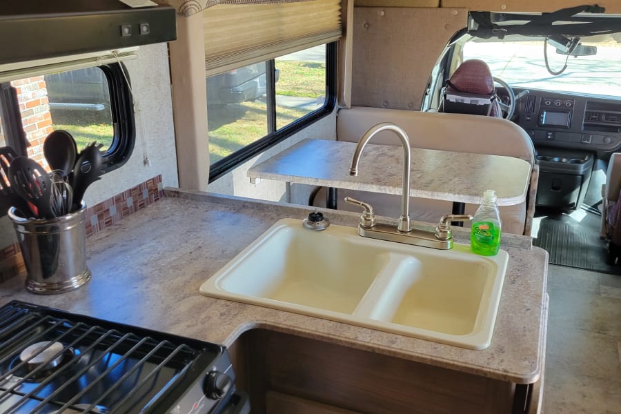 Inside this RV rental: a compact kitchen with stove, sink, microwave, and dinette. Driver's area visible beyond.
