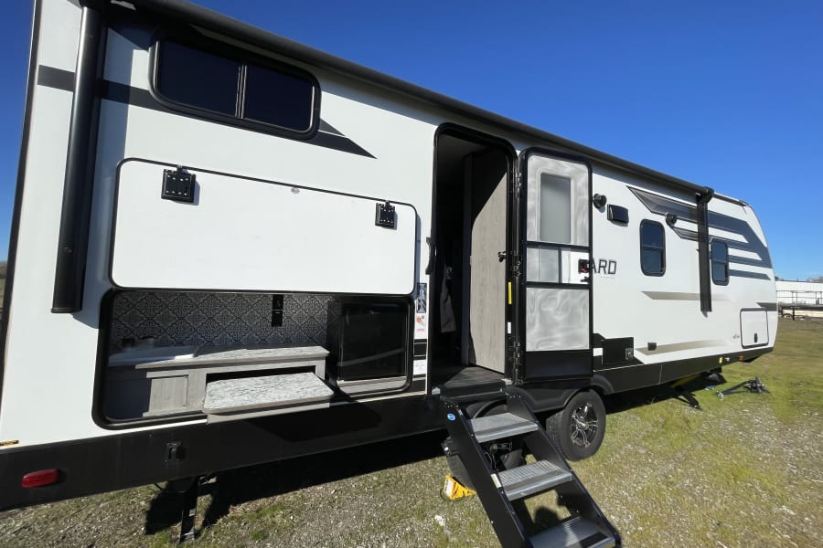 Explore this white and black RV rental with its convenient outdoor kitchen setup and sturdy entry steps, ready for your next adventure.