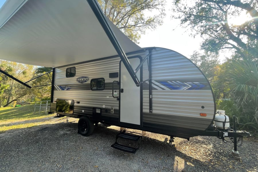Explore the outdoors! This RV rental, a travel trailer with an extended awning, sits on a gravel lot under sunny trees.