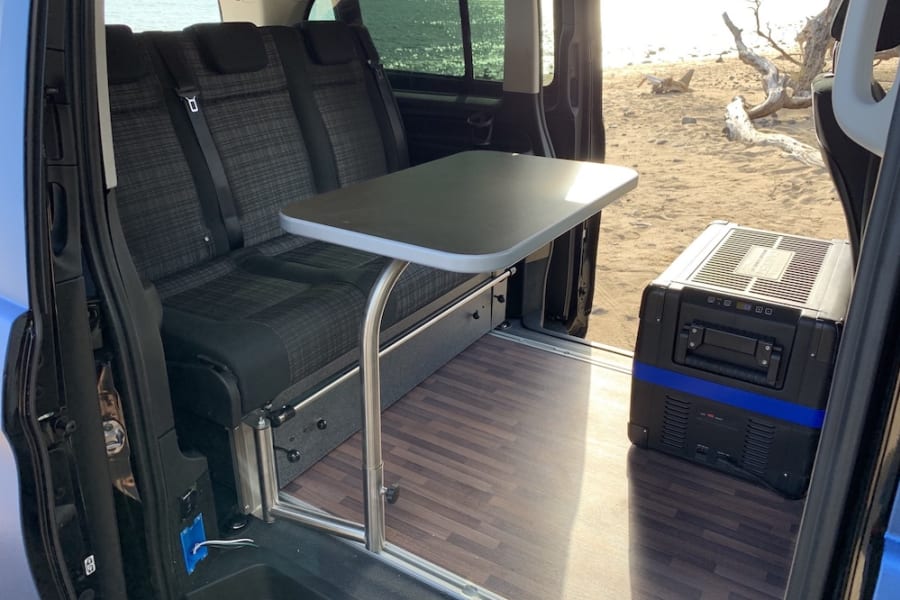 RV rental's versatile interior: seating, table, and portable cooler, with an open door view of a sunny beach.