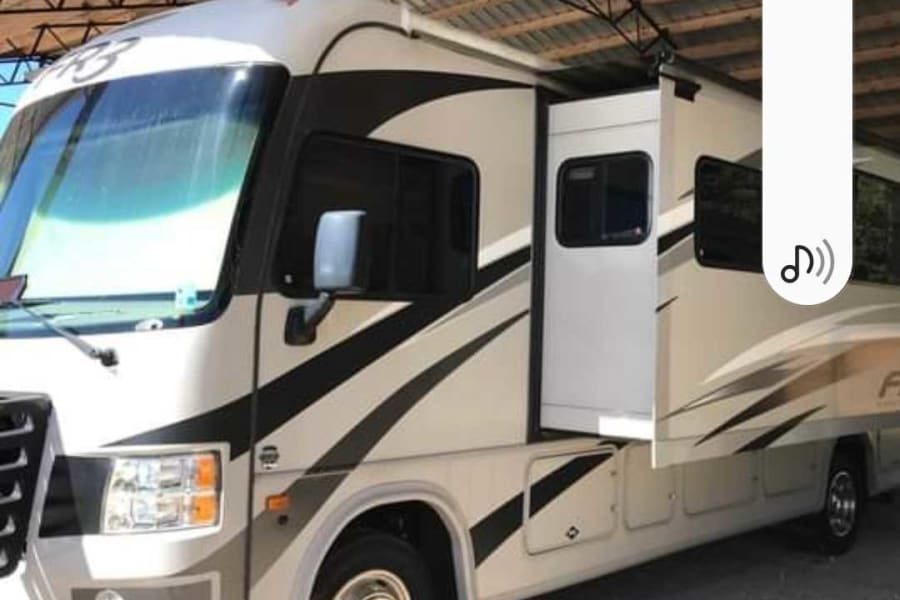Modern RV rental, light-colored with dark accents, slide-out extended, parked under a sturdy shelter.