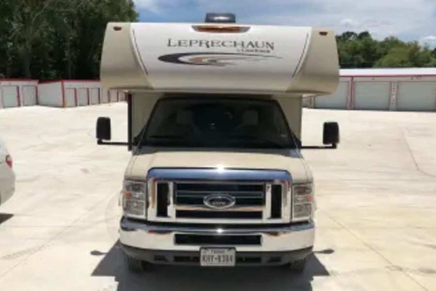 Front view of a tan Ford Leprechaun RV rental, ready for travel. Parked on a paved lot with storage units.