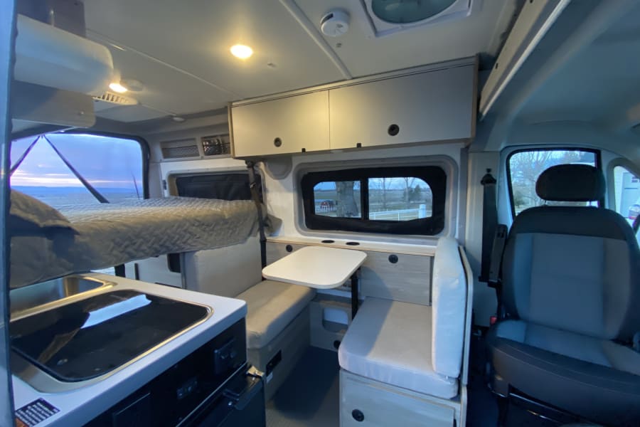 Interior of an RV rental camper van, featuring a bed, kitchenette, and dining area under warm lighting.