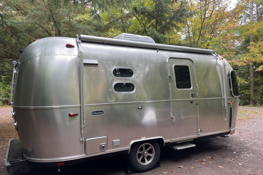 A silver Airstream Caravel travel trailer, glistening from rain at a fall campsite. Perfect rv rental for your outdoor adventure!