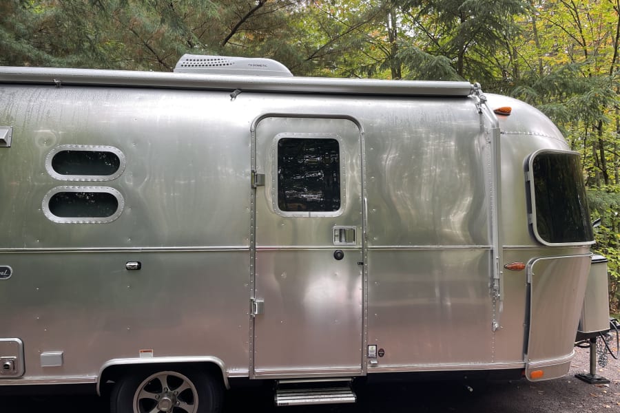 A shiny silver Airstream RV rental gleams under green trees, showing its side profile with windows and door.