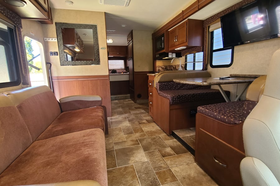 RV rental interior: living area with sofa, dining booth, kitchen with sink/microwave, and TV. Tile flooring visible.