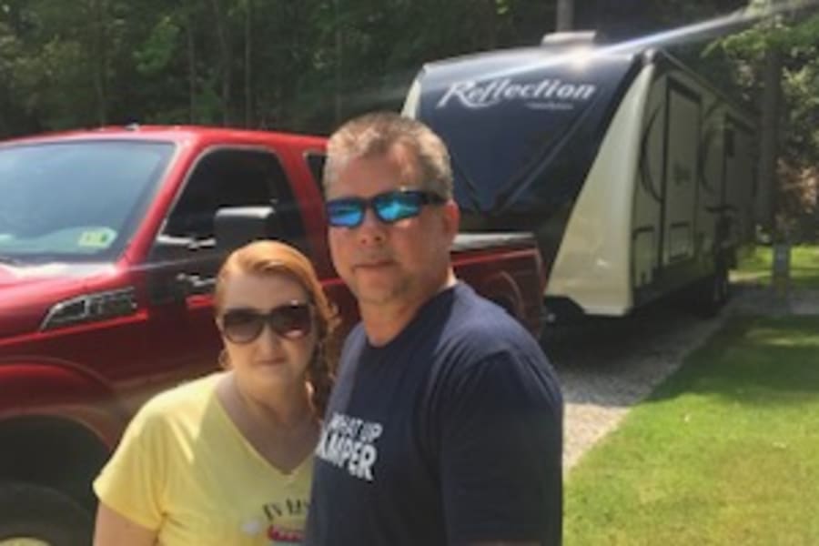 A couple with their large red truck & towed white fifth-wheel RV at a sunny campsite. Perfect for your next RV rental adventure!