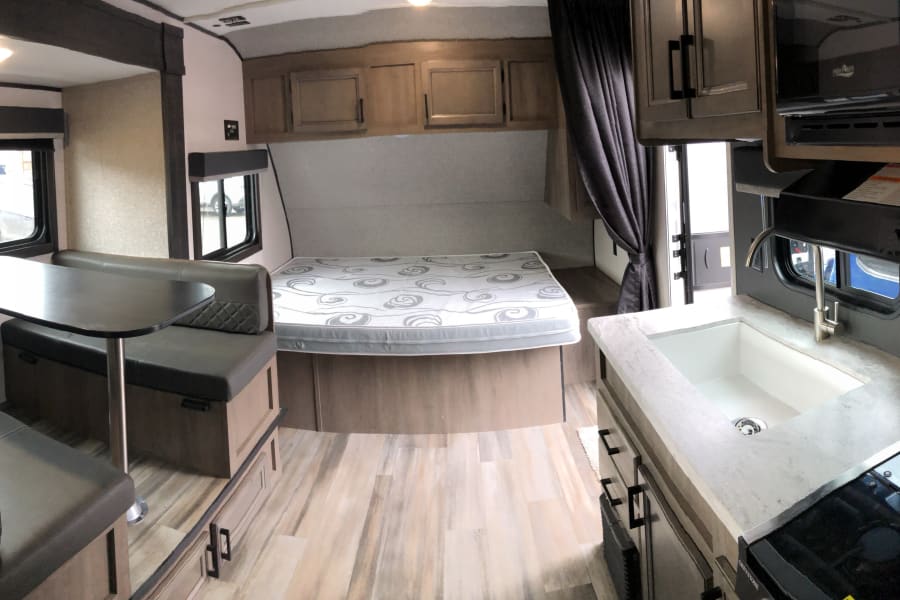 An RV rental interior with a dinette, bed, and a compact kitchen featuring a sink, stovetop, and microwave.