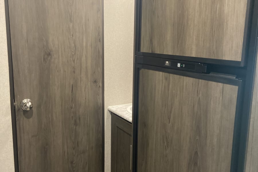 An RV rental interior with a wood-look refrigerator and a wooden door, hinting at a compact bathroom beyond.