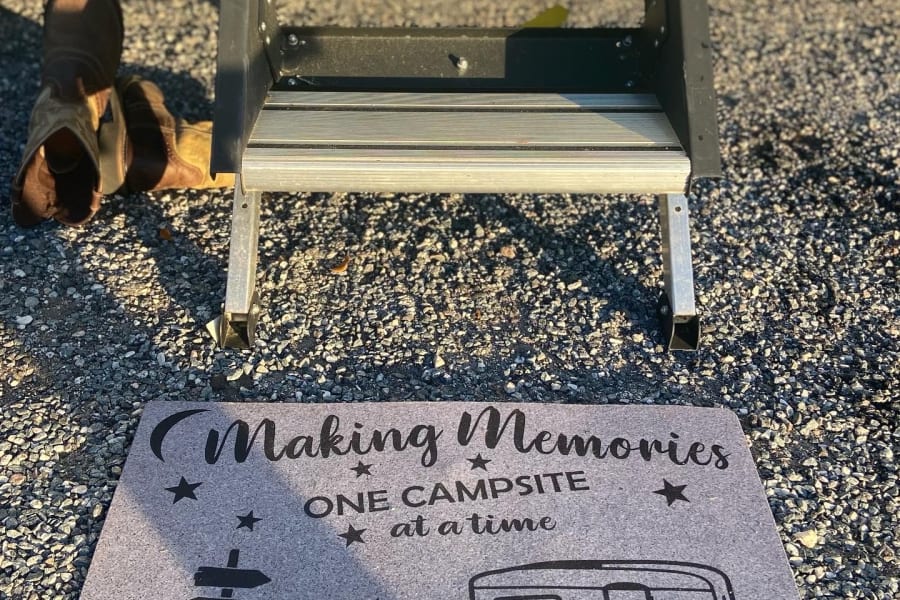 RV rental steps & doormat on gravel. Custom mat reads 'Making Memories ONE CAMPSITE at a time'. Camping boots nearby.
