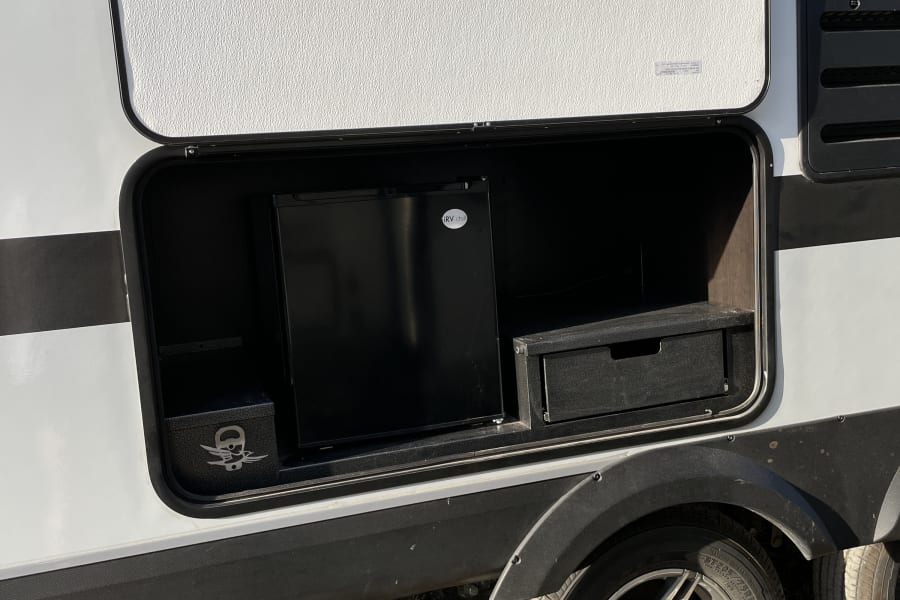 Open compartment on this rv rental reveals an 'RV Chill' mini-fridge and a storage box, offering convenient outdoor amenities. Jayco RV on grass.