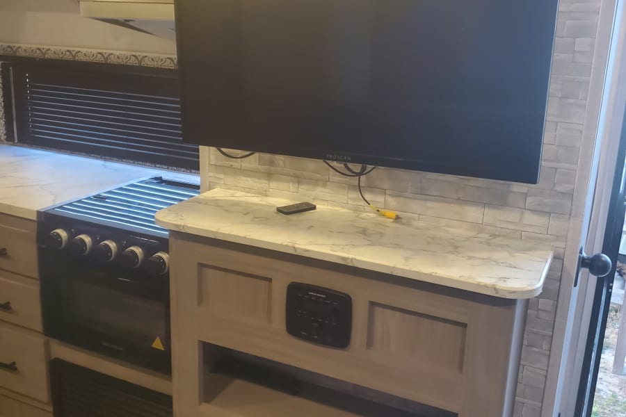 Interior of an RV rental, showing a kitchen with a stove, microwave, and a living area with a TV and electric fireplace.