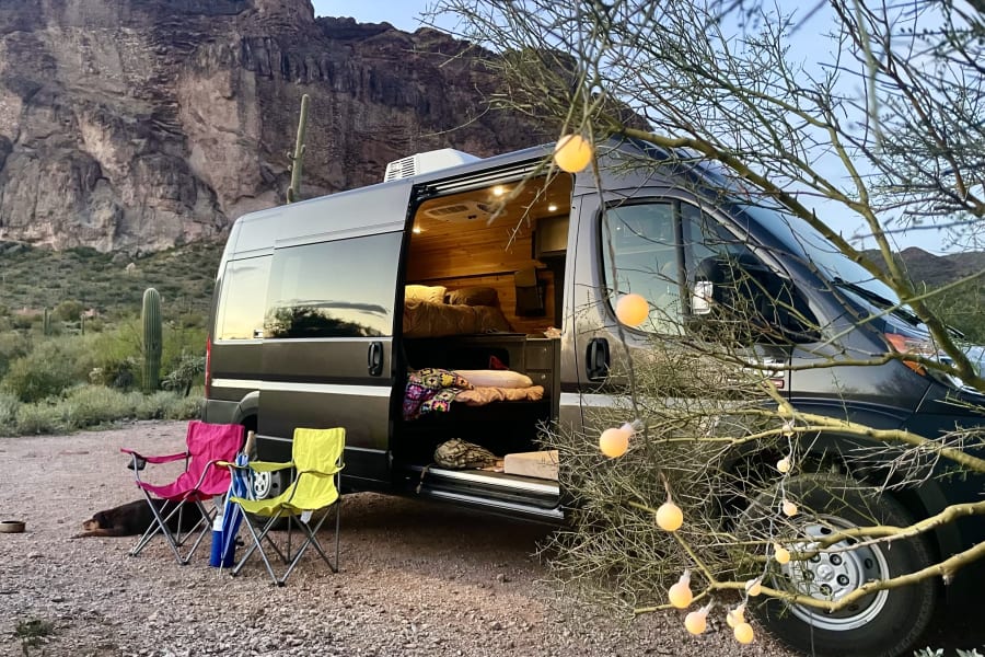 Cozy RV rental camper van parked in the desert with mountains. Open door reveals interior, chairs out, dog sleeping, string lights glow.