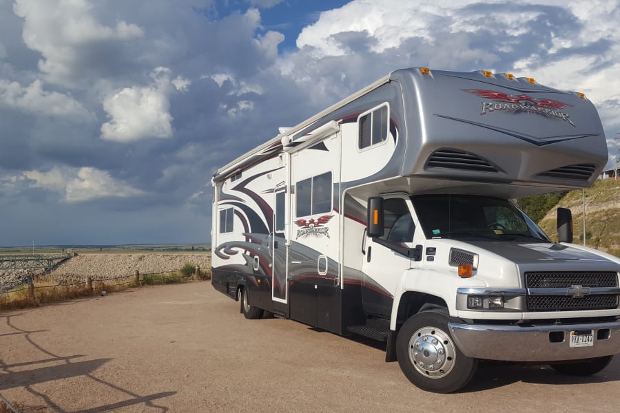 White & black Road Warrior RV rental, a large motorhome, parked on a dirt lot under cloudy skies. Bridge visible.