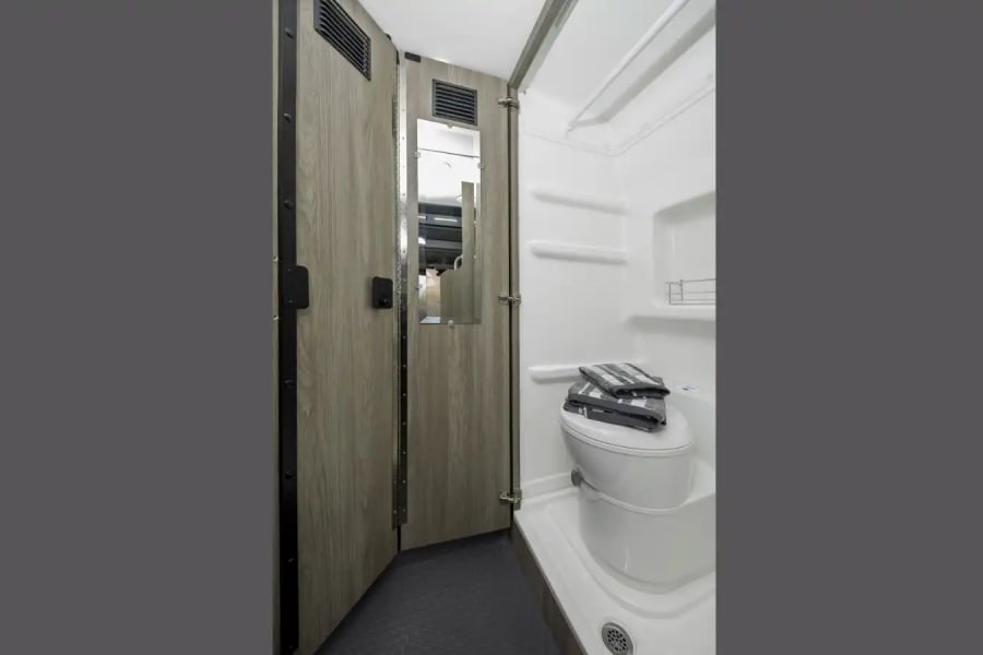 RV rental bathroom: compact white shower, toilet with towels, and wood-paneled door with mirror.