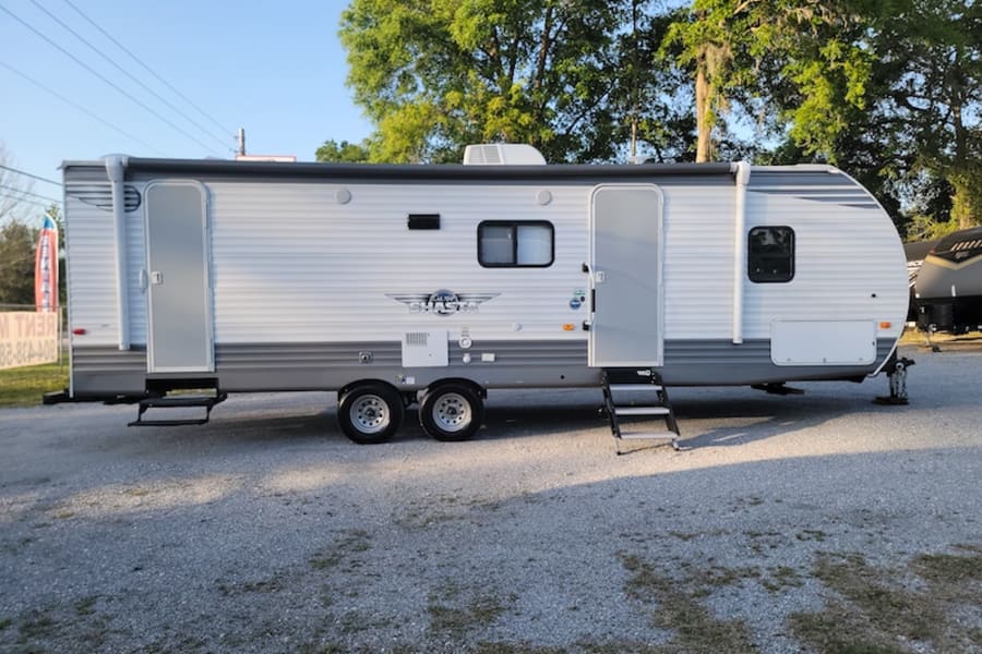 White and gray travel trailer RV rental with two doors and steps, parked on a gravel lot, ready for your next adventure.