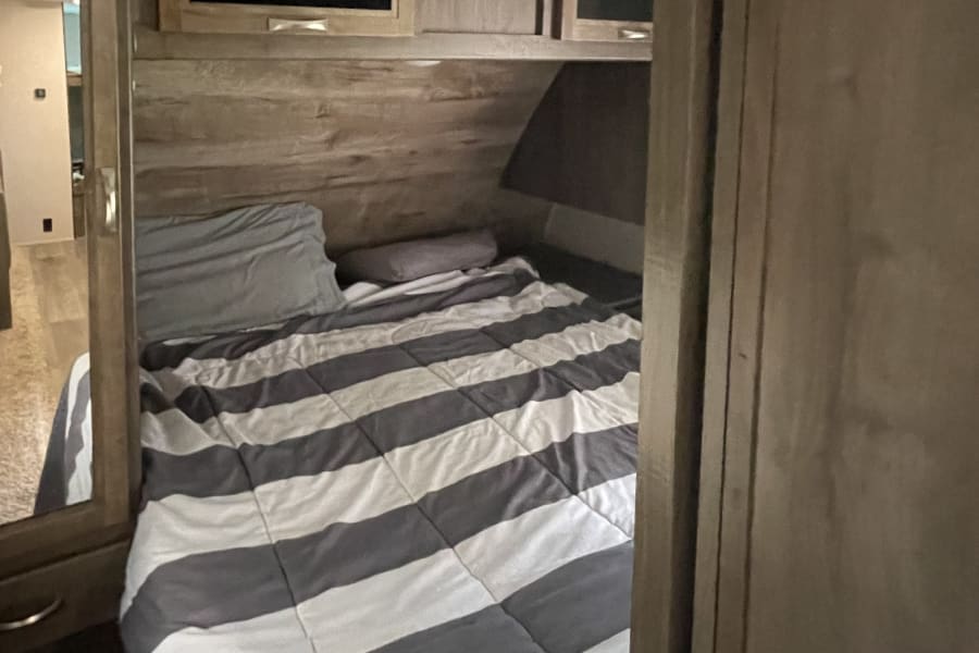 Comfortable RV rental bedroom featuring a striped bed, mounted TV, and overhead storage for your road trip comfort.