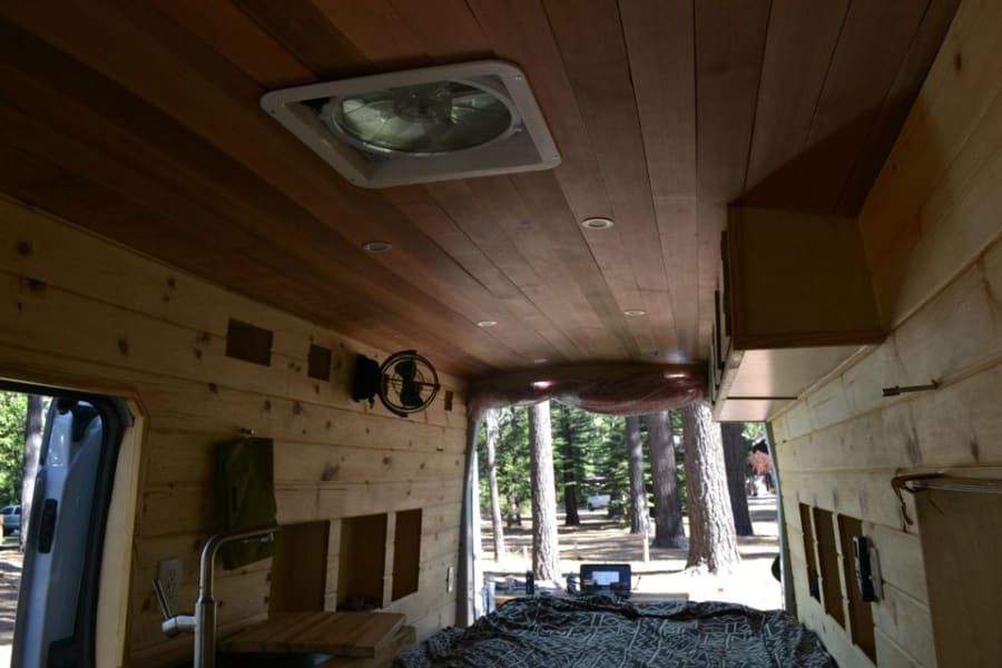 Cozy RV rental interior with wooden ceiling, vent fan, and bed. Includes a small kitchen sink and a clear view of nature.