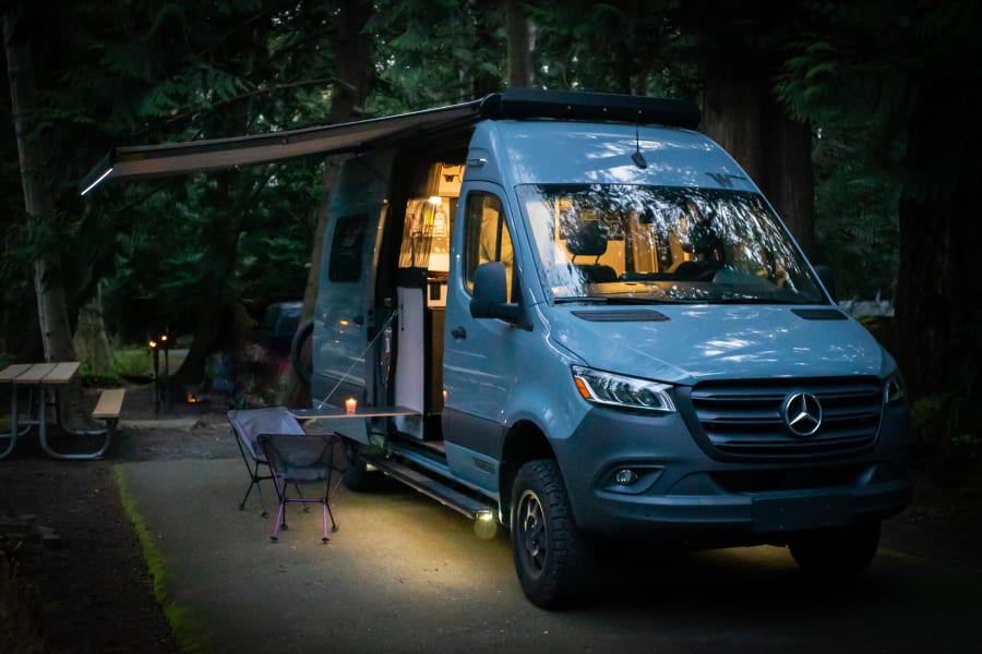 An illuminated gray Mercedes-Benz Sprinter van RV rental parked at a serene forest campsite at dusk, with an awning and outdoor seating.